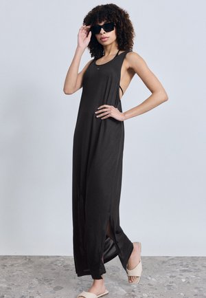 Femeie cu păr creț, îmbrăcată într-o rochie maxi neagră fără mâneci, ochelari de soare și sandale beige, stând pe podea gri, în fața unui perete simplu.