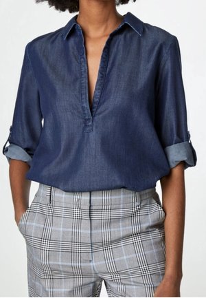 Femme portant un chemisier bleu marine à manches longues avec manches retroussées et un pantalon taille haute à carreaux gris, mains dans les poches.