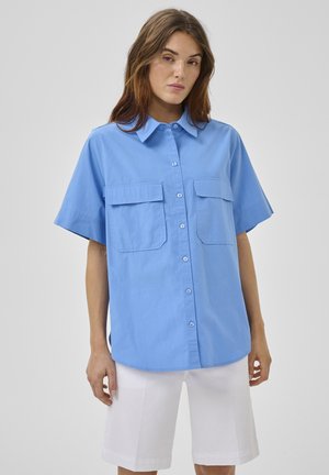Vrouw draagt een lichtblauw overhemd met korte mouwen en knopen, met borstzakken, en witte knielange shorts tegen een grijze achtergrond.