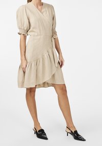 Beiges Leinen-Wickelkleid mit Puffärmeln, Rüsche am Saum und verstellbarem Bindegürtel. Kombiniert mit schwarzen Slingbacks mit spitzer Zehenpartie.