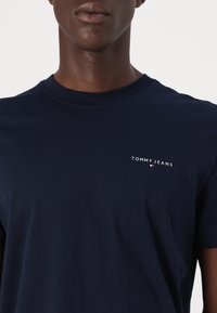 Granatowy bawełniany t-shirt z okrągłym dekoltem i krótkimi rękawami. Na lewej piersi znajduje się biały napis „TOMMY JEANS”. Gładka faktura.