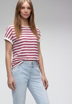 Jeune femme aux longs cheveux blonds portant un t-shirt rayé violet et blanc et un jean bleu clair, debout devant un fond gris uni.