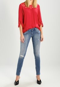 Blusa rossa con dettagli in pizzo al collo e alle maniche, abbinata a jeans blu strappati e décolleté neri a punta.