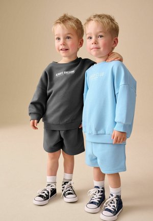 Kaksi nuorta poikaa seisoo vierekkäin, toinen harmaassa collegepaidassa ja shortseissa, toinen vaaleansinisessä collegepaidassa ja shortseissa, molemmilla tennarit jalassa.