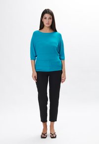 Maglione turchese con texture a zigzag, maniche a tre quarti, abbinato a pantaloni neri aderenti e sandali. Sfondo bianco semplice.