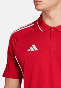 Rode polo-shirt met witte kraag en accenten. Bevat drie witte strepen op de schouders. Merkmening geborduurd op de linkerkant van de borst.