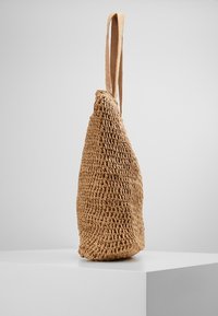 Vävd halmtoteväska i ljusbrun färg med en tårform och två tygaxelremmar. Texturerad utsida med öppen vävstruktur.