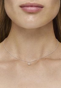 Collar de cadena de plata que presenta un pequeño colgante redondo, exhibido contra un escote liso y sin adornos y una textura de piel sutil.