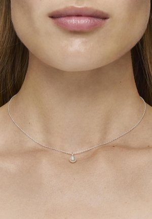Collar de cadena de plata que presenta un pequeño colgante redondo, exhibido contra un escote liso y sin adornos y una textura de piel sutil.