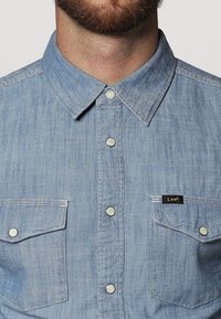 Vaaleansininen denimpaita, jossa on klassinen kaulus, napitus edessä ja kaksi rintataskua. Varustettu pienellä mustalla "Lee"-merkillä. Pehmeä rakenne.