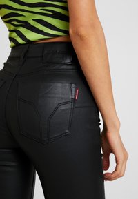 Svarta imitationläderbyxor med en åtsittande design, med två bakfickor och en liten röd märkesskylt på höger ficka. Slät textur.