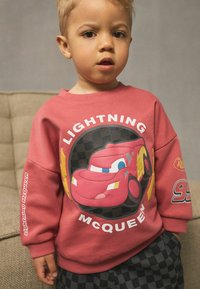 Un tout-petit portant un sweat-shirt rouge de Lightning McQueen et un pantalon à carreaux noirs, debout à l'intérieur près d'un canapé beige.