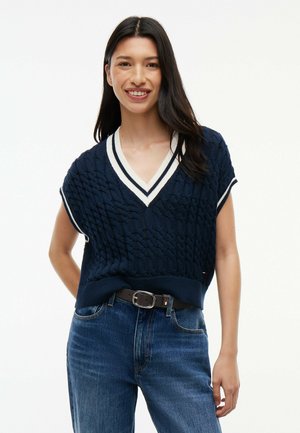 Ung kvinde med langt mørkt hår smiler iført en mørkeblå kabelstrikket ærmeløs sweater med hvidstribet V-hals og blå jeans.