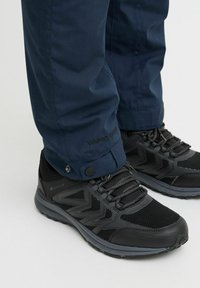 Pantofi sport negri cu plasă texturată și tălpi din cauciuc, asortați cu pantaloni impermeabili de culoare bleumarin, care au închideri cu capse și branding "W-PRO 10000".