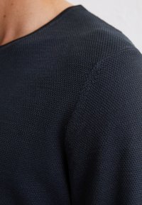 Granatowy, dzianinowy sweter z teksturowanym wykończeniem i okrągłym dekoltem, uchwycony z okolic ramienia i szyi.