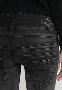 Jeans de mezclilla negra con un diseño ajustado, con bolsillos traseros cosidos y una etiqueta de cuero en la pretina. Tejido texturizado con detalles desgastados.