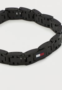 Βραχιόλι με μαύρες μεταλλικές συνδέσεις και ορθογώνια τμήματα, με ένα μικρό έμβλημα σημαιάκι σε κόκκινο, λευκό και μπλε σε μία σύνδεση.