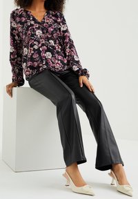 Žena v černé květinové blouse, černých kožených kalhotách a bílých slingback podpatcích, sedící na bílém krychlovém stole na neutrálním pozadí.