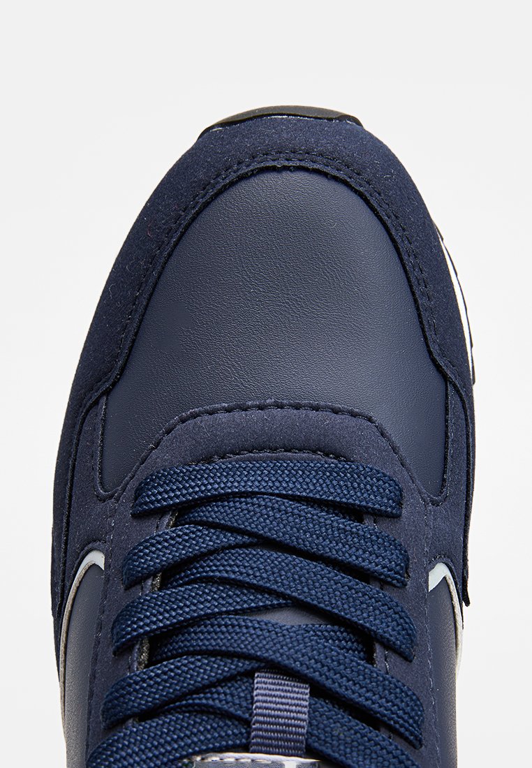 Zapatilla de deporte azul marino con parte superior de cuero y ante, puntera redonda, cordones planos y cuello acolchado. Presenta un acento de logo blanco en el lateral.