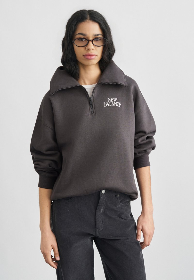 Φούτερ με φερμουάρ από ανθρακί ύφασμα, με ψηλό γιακά και κεντημένο λογότυπο "NEW BALANCE" στο αριστερό μέρος του στήθους, σε συνδυασμό με παντελόνι σκούρου γκρι χρώματος.