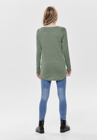 Långärmad olivgrön topp med rundad fåll, kombinerad med blå skinny jeans och svarta ankelkängor. Slät textur och åtsittande design.