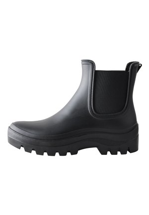 Person wearing black ankle rain boots, gray socks, and a brown skirt standing on green grass outdoors.