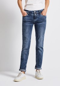 Jeans de mezclilla de altura media en un lavado azul deslavado. Cuenta con parches desgastados, cinco bolsillos y dobladillos enrollados. Combinados con zapatillas blancas.