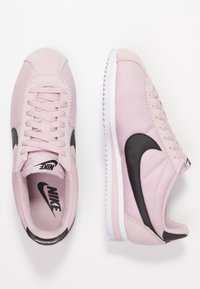 Ett par ljusrosa Nike-sneakers med svarta swoosh-logotyper, vita sulor och snören på en vit bakgrund, där den ena visar ovanifrån och den andra från sidan.