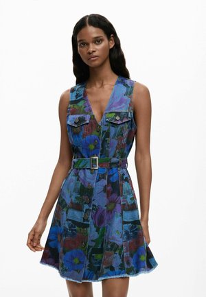 Kvinde iført ærmeløs denimkjole med knapper foran, dekoreret med flerfarvede blomster- og grafiske prints, med bælte i taljen, stående mod en hvid baggrund.