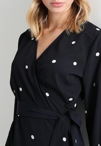 Robe portefeuille marine en tissu léger à pois blancs, avec un profond décolleté en V, des manches longues et une ceinture assortie à nouer.