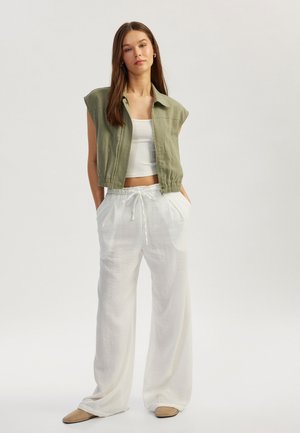 Chaqueta verde oliva corta con mangas cortas, combinada con pantalones de lino blancos de corte suelto y zapatos beige de tipo slip-on. Texturas lisas en toda la prenda.