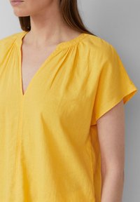 Haut jaune à manches courtes avec un décolleté en V profond, fabriqué en tissu léger. Détails froncés aux épaules, texture lisse, coupe décontractée.
