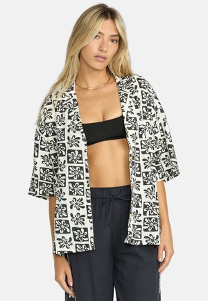 Camicia a maniche corte in tessuto bianco con un motivo floreale nero. Vestibilità ampia con una parte anteriore aperta e trame a contrasto.