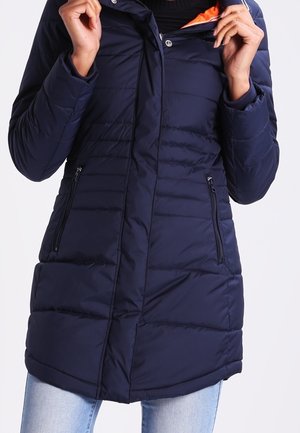 Personne portant une veste d'hiver matelassée bleu marine avec des poches zippées, tenant le col partiellement dézippé qui révèle une doublure intérieure orange.