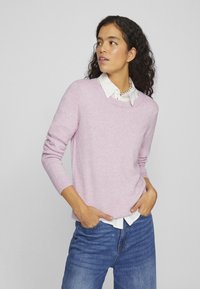 Light pink sweater with crew neck, ribbed cuffs, and a relaxed fit, layered over a white collared shirt. Denim jeans complete the outfit.