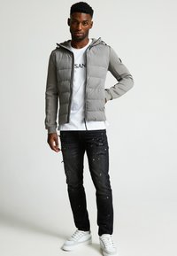 Giacca imbottita grigia con cappuccio, indossata su una t-shirt bianca. Abbinata a jeans neri strappati e sneaker bianche.