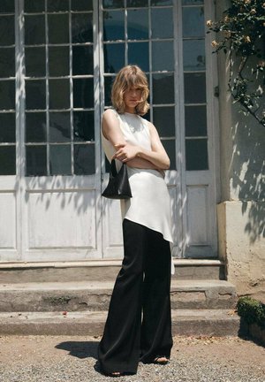 Un haut sans manches blanc avec un ourlet asymétrique associé à un large pantalon noir. Un petit sac à main noir complète la tenue. Fond en pierre.