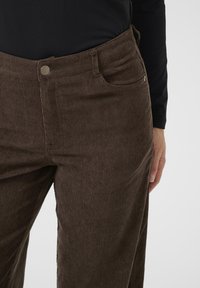 Pantalons en velours côtelé marron avec une surface texturée, dotés d'une poche avant, de passants de ceinture et d'une fermeture à bouton en métal.