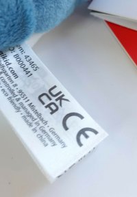 White care label with black text showing UKCA, CE markings, product details, and manufacturer information, attached to plush blue material.