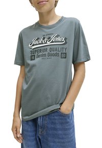 Camiseta de algodón gris con mangas cortas que presenta un logo impreso "Jack & Jones" y "Superior Quality Denim Goods" en blanco y negro.