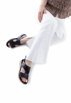 Sandali in pelle nera traforata con punta e lati aperti, dotati di suola zeppa in legno chiaro e dettaglio annodato sul retro.