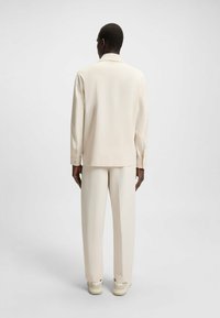 Giacca beige chiaro con una vestibilità rilassata e maniche lunghe, abbinata a pantaloni larghi coordinati. Texture liscia e design minimal.