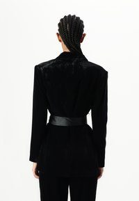 Black velvet blazer with a shawl collar and long sleeves, cinched at the waist with a wide black leather belt. Smooth texture, tailored fit.