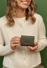 Vihreä nahkainen lompakko, kompakti muotoilu, ommellut reunat, vetoketjusuljenta, kohokuvioitu brändilogo, pidetään käsissä, yhtenäistä vihreää taustaa vasten.