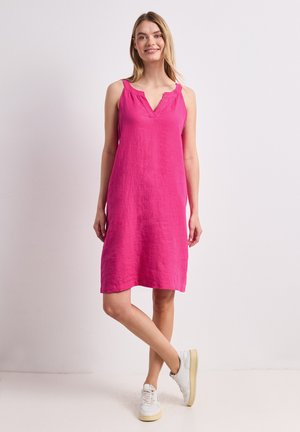 Femme debout, jambes croisées, portant une robe sans manches rose vif longueur genou et des baskets blanches sur un fond uni.