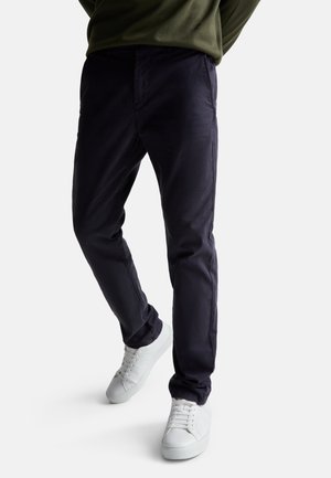 Persona con pantalones azul marino oscuro y zapatillas blancas, de pie contra un fondo claro y liso.
