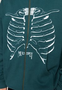 Sudadera color azul verdoso con un gran gráfico de un esqueleto de tórax blanco en la parte delantera, con cierre de cremallera y dos bolsillos frontales.