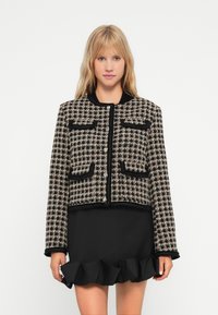 Tweed kort jakke i svart og krem med et geometrisk mønster, som har fire frontlommer og rund hals. Stylet med et volangminiskjørt i svart.