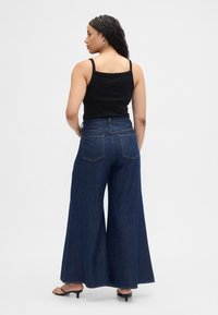 Široke kavbojke z visokim pasom v temno modri barvi, s petimi žepi in kontrastnim šivom. Doplačane s črno rebrasto majico brez rokavov.