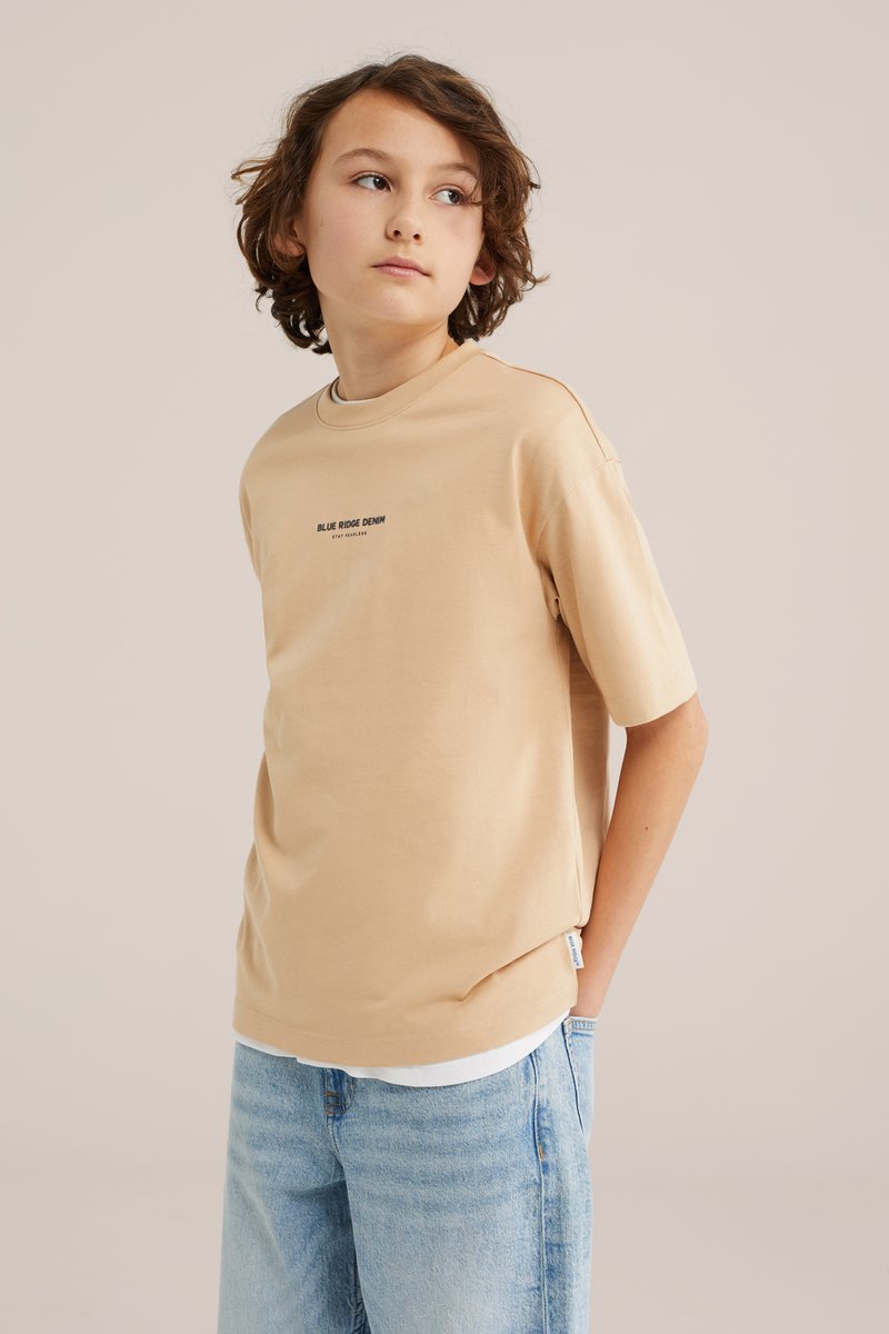 Jeune personne aux cheveux bouclés portant un T-shirt beige "Blue Ridge Denim" et un jean bleu clair, regardant sur le côté sur un fond uni.